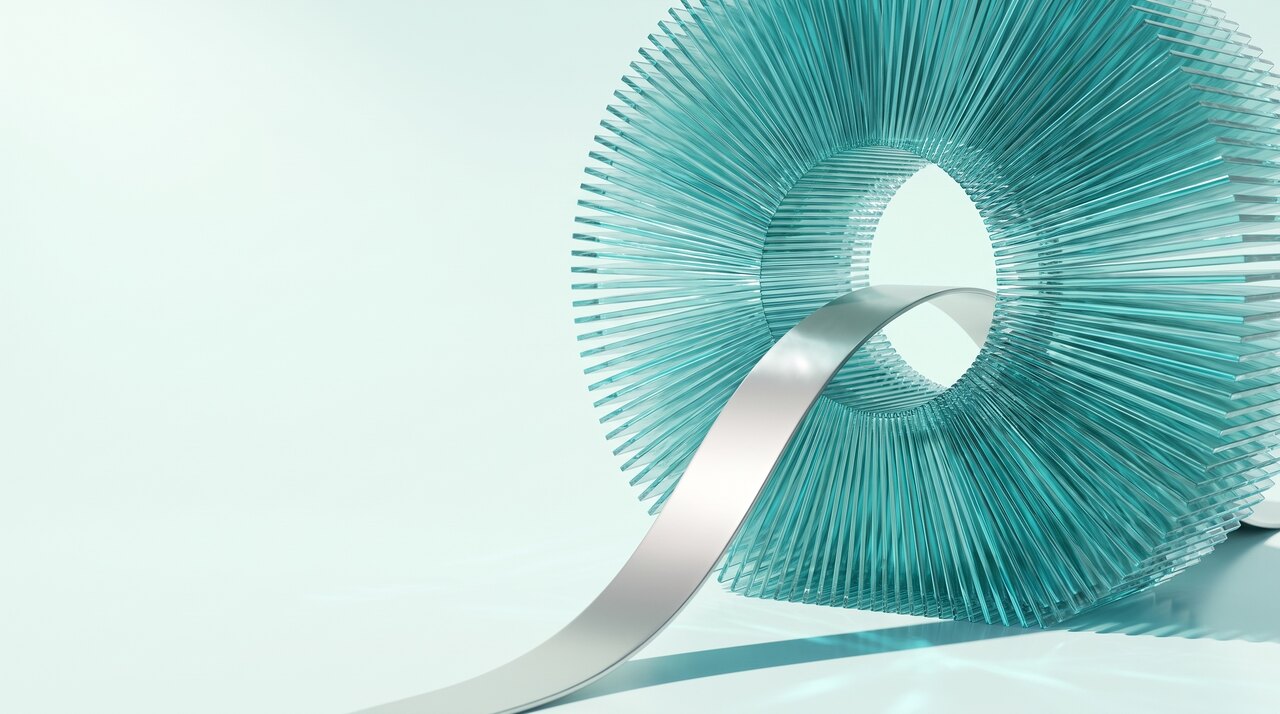 A material study in bright daylight. A dense radial wheel of clear turquoise glass blades with one clear opening at its centre, and a single pearl silver ribbon curving down and away from that opening across the pale floor.